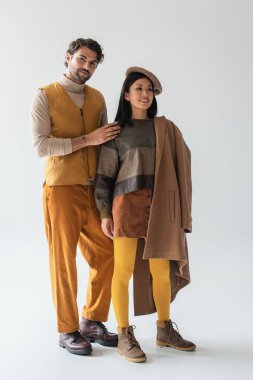 Asyalı, bereli ve gri taytlı bir kadının yanındaki kameraya bakan trend bir adam.