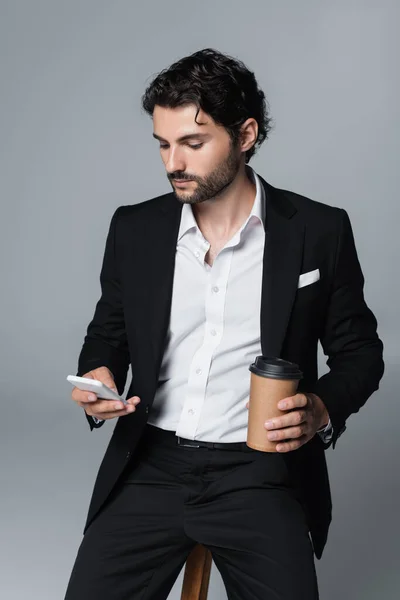 穿着时髦的黑发男人 一边喝着咖啡一边用手机 一边孤零零地坐在灰色的椅子上 — 图库照片