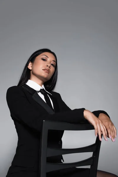 官能的なアジアの女性で黒ブレザーとタイポーズオン椅子絶縁グレー — ストック写真