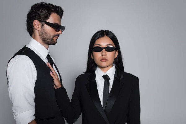 elegant asian woman in black formal wear and sunglasses looking at camera near stylish man isolated on grey