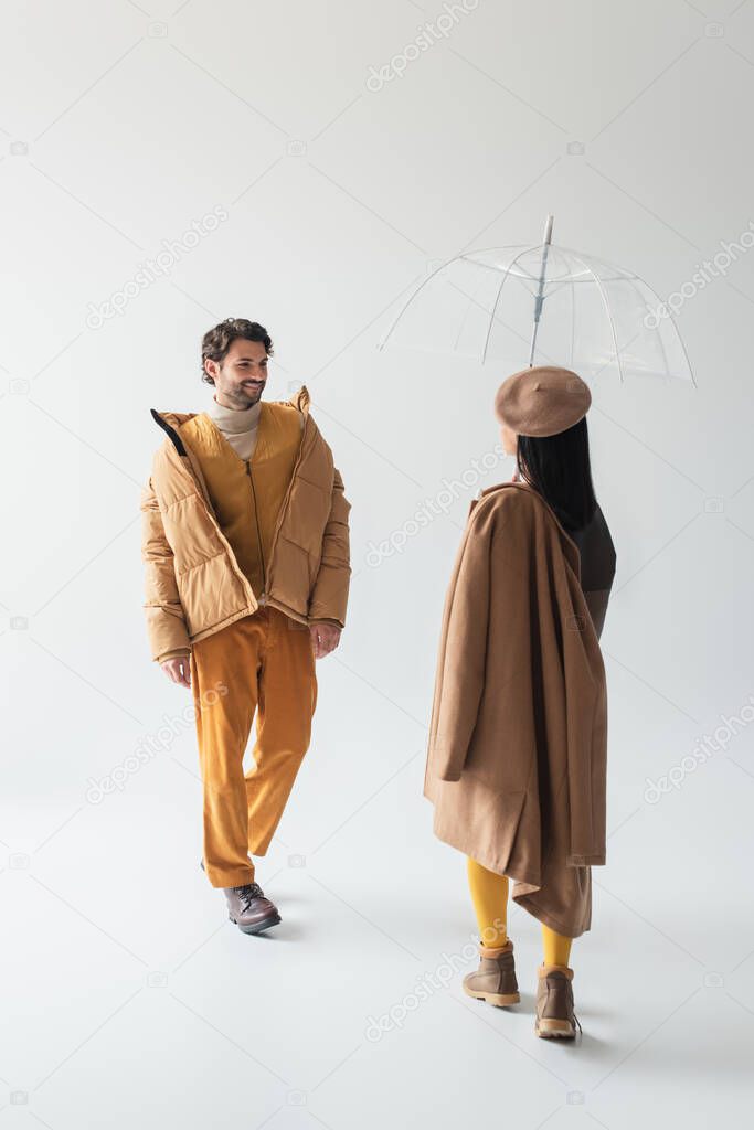 woman in transparent umbrella and smiling man in beige puffer jacket walking towards each other on grey