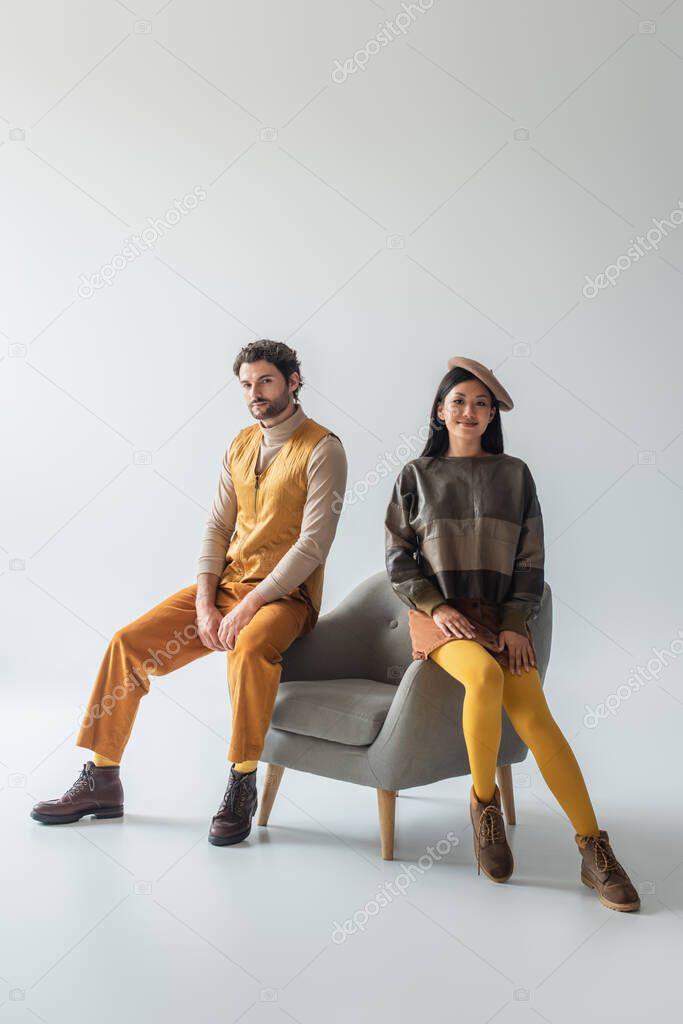 multiethnic couple in fashionable clothes sitting on armchair and looking at camera on grey