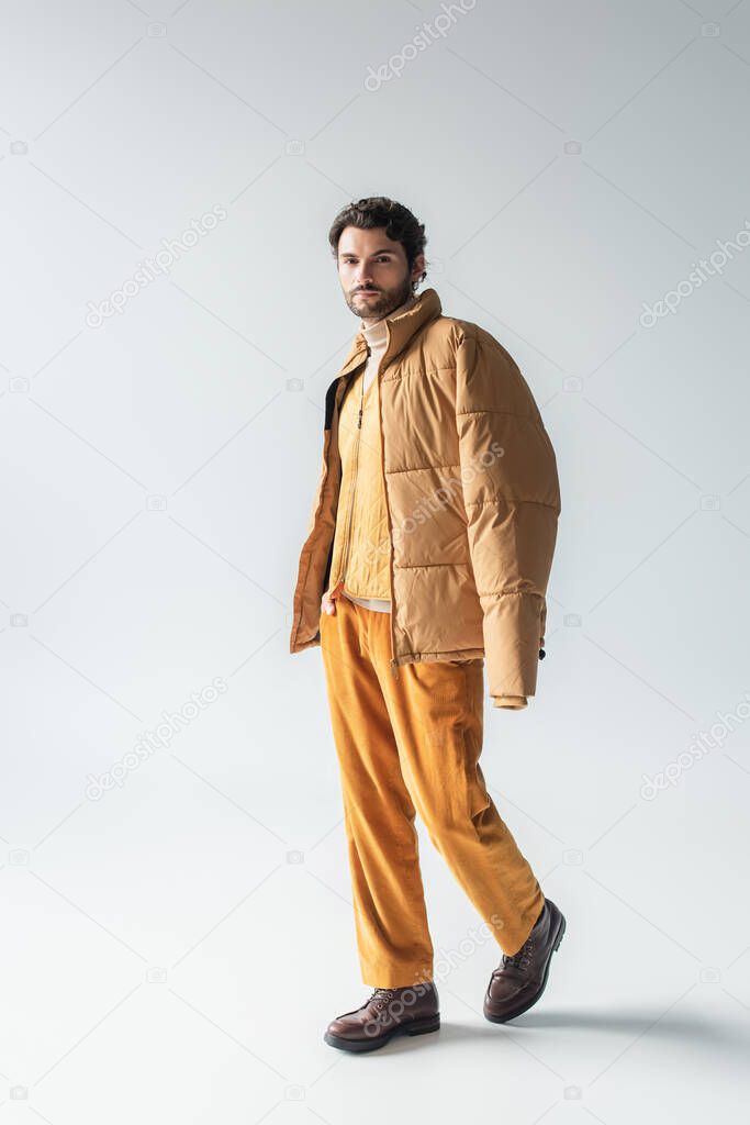 full length view of man in warm puffer jacket and yellow trousers posing with hand in pocket on grey