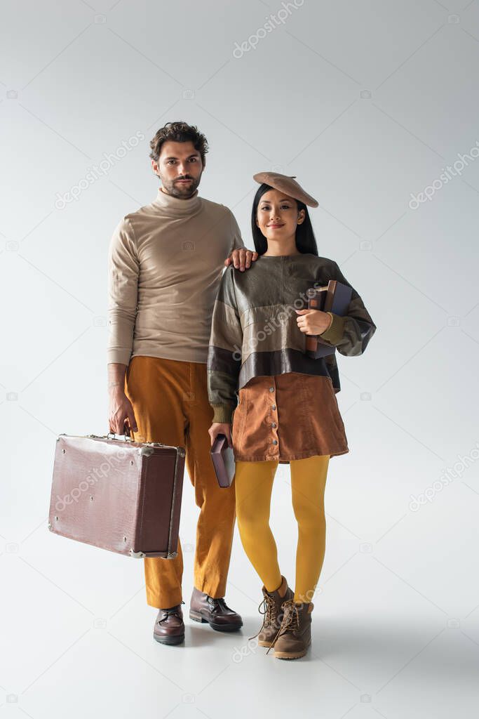 full length view of asian woman with books and stylish man with vintage suitcase on grey