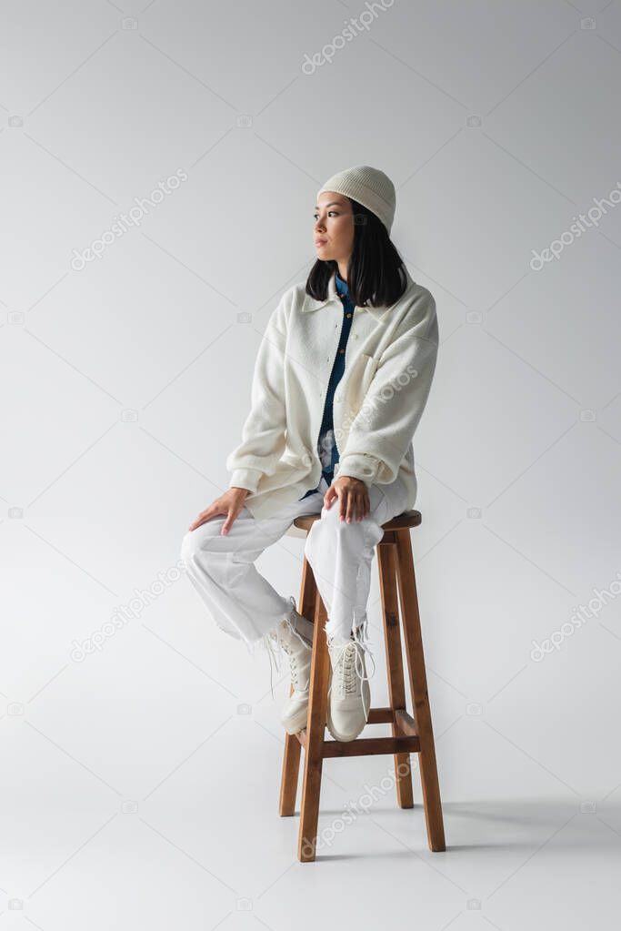 full length view of trendy asian woman in white clothing sitting on high stool on grey