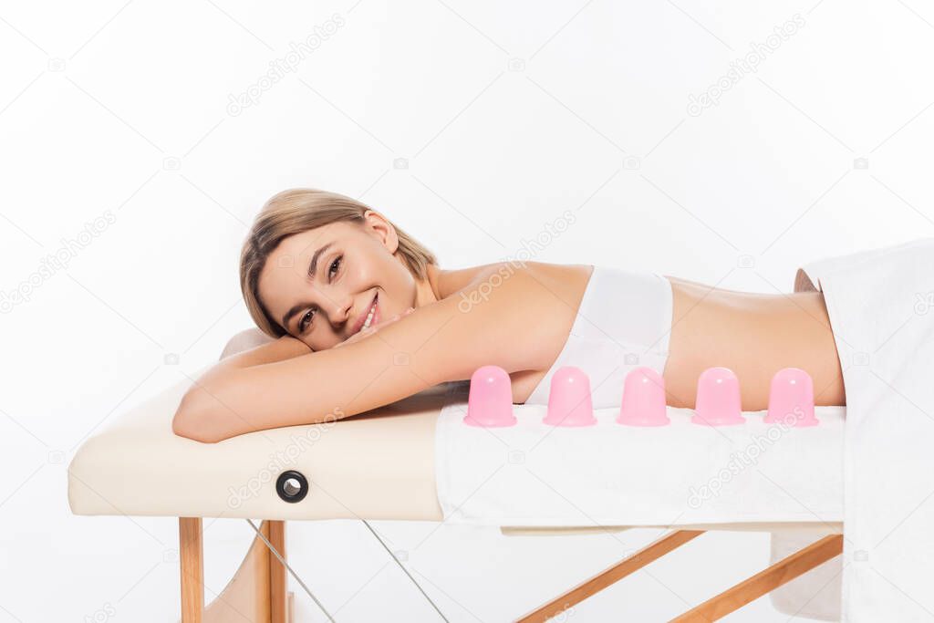 happy young woman lying on massage table near pink massage cups isolated on white