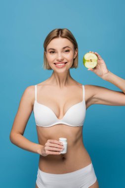 Beyaz iç çamaşırlı neşeli bir kadın elinde vitaminli bir şişe ve yarı izole bir elma tutuyor. 