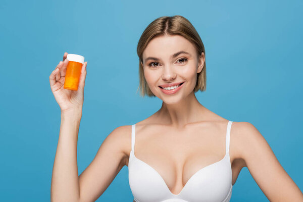 happy young woman in white bra holding bottle with vitamins isolated on blue 
