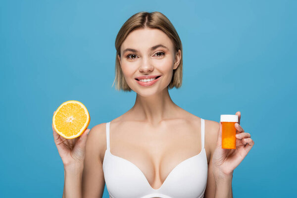 smiling young woman in white lingerie holding bottle with vitamins and orange half isolated on blue 