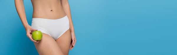 обрезанный вид молодой женщины в белом нижнем белье держа зеленое яблоко изолированы на синий, баннер