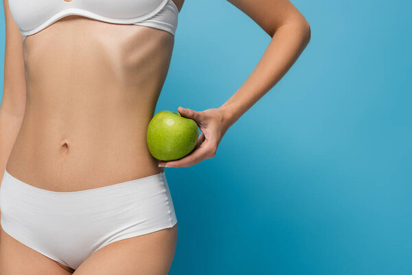 частичный взгляд на стройную молодую женщину в нижнем белье, держащую яблоко изолированы на синий 
