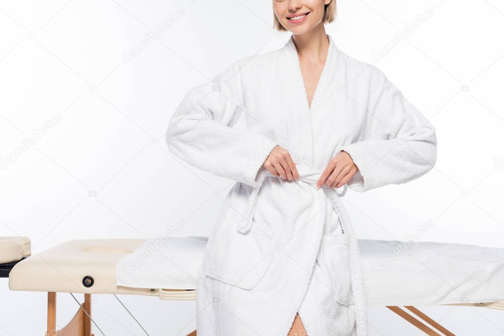 cropped view of young cheerful woman in bathrobe near massage table on white