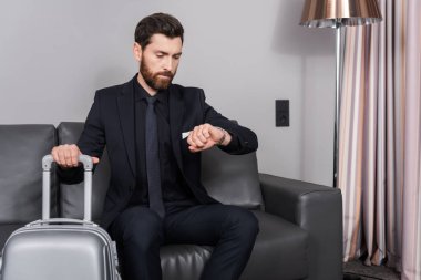 Sakallı iş adamı kol saatini kontrol ediyor ve otel odasında bagajın yanında oturuyor.
