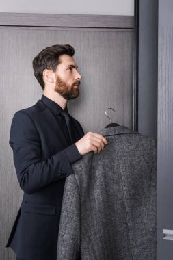 Otel odasındaki dolabın yanında askı tutan takım elbiseli sakallı adamın yan görüntüsü. 