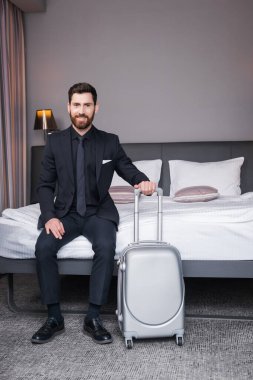 Takım elbiseli neşeli bir adam elinde bavul tutarak otel odasında yatakta oturuyor. 