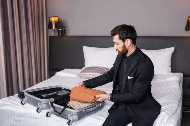 Otel odasında bavulunu boşaltan takım elbiseli sakallı bir adam. 