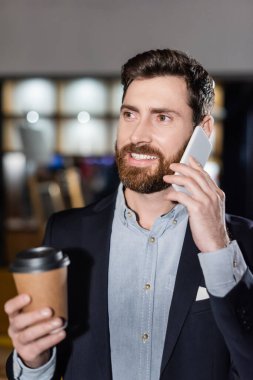 Takım elbiseli neşeli adam akıllı telefonla konuşuyor ve otel lobisinde bulanık kağıt bardak tutuyor.