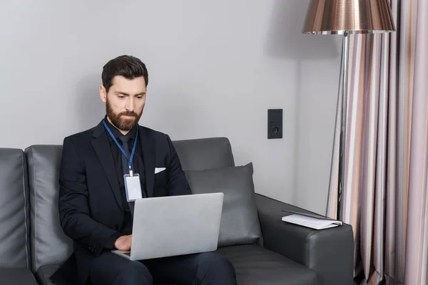ホテルの部屋にある革張りのソファに座っている間にノートパソコンを使ったIdバッジを持つ髭のビジネスマン — ストック写真