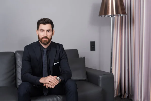 bearded businessman in suit sitting on leather couch in hotel room
