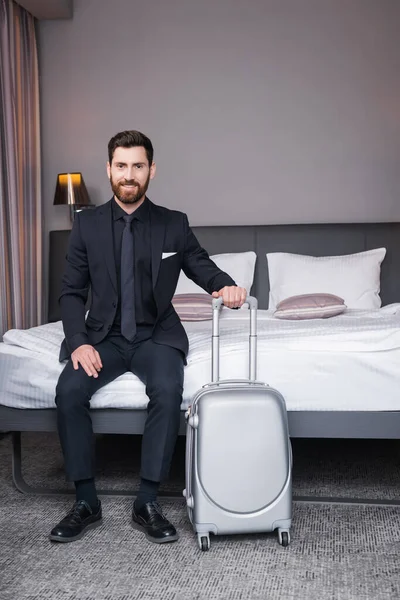 快乐的穿西装的男人拿着行李把手坐在旅馆房间的床上 — 图库照片