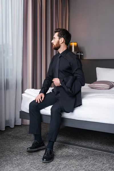 bearded man in eyeglasses and suit sitting on bed in hotel room