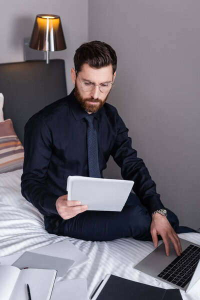 bearded man in suit and eyeglasses looking at folder and using laptop in hotel