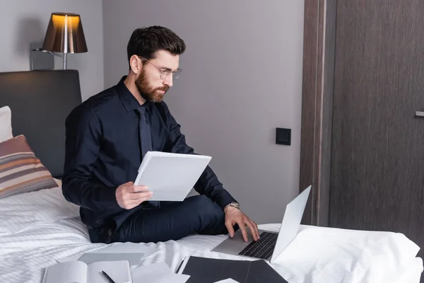 Skäggig Man Kostym Och Glasögon Med Laptop Nära Mappar Hotellsäng — Stockfoto