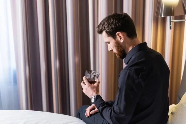 满脸胡须的男人举着威士忌 坐在旅馆的床上 — 图库照片