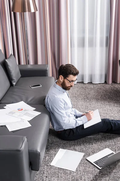 戴眼镜的大胡子男子坐在地毯上 在笔记本电脑和沙发旁边的笔记本上写字 — 图库照片