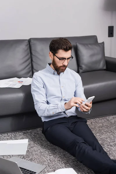 Freelance Barbuto Occhiali Seduti Moquette Utilizzando Smartphone Vicino Laptop Divano — Foto Stock