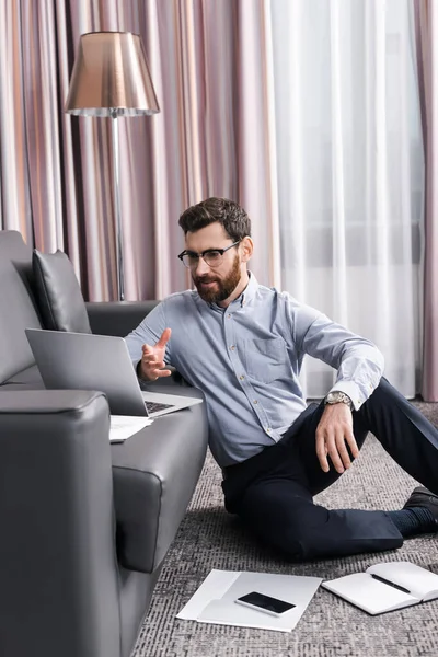 カーペットの上に座ってノートパソコンでビデオ通話中に話す眼鏡の髭の男 — ストック写真