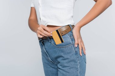 cropped view of young woman putting credit card with cashback in pocket on jeans isolated on grey clipart