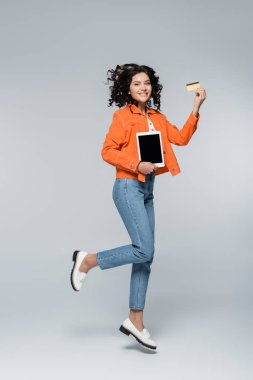 Turuncu ceketli, elinde nakit para ve dijital tabletli kredi kartı olan mutlu bir kadın.