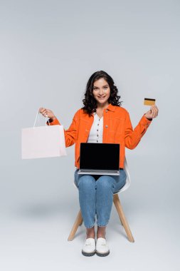 Neşeli kadın alışveriş çantası ve kredi kartını gri ekranlı dizüstü bilgisayarın yanında tutuyor. 
