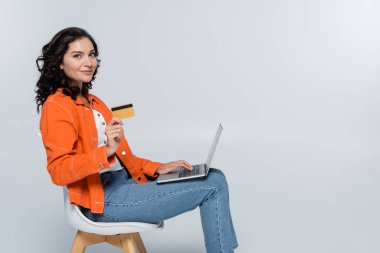 Laptop kullanan turuncu ceketli ve kredi kartını gri renkte tutan memnun bir kadın. 