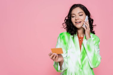Kravatlı, ceketli, elinde kredi kartı olan mutlu genç bir kadın pembe bir telefondan izole bir şekilde konuşuyor. 