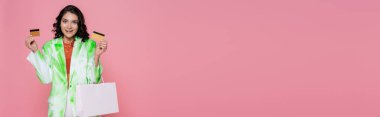 Pembe pankartta izole edilmiş, elinde kredi kartı ve alışveriş çantası tutan kravatlı genç bir kadın.
