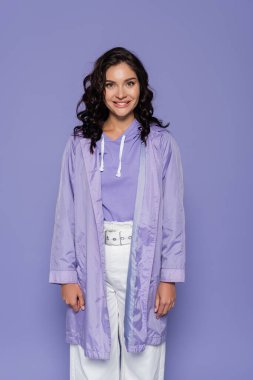 happy young woman in raincoat smiling isolated on purple  clipart