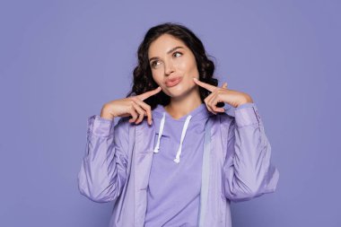 brunette young woman in raincoat pouting lips and pointing at cheeks isolated on purple clipart