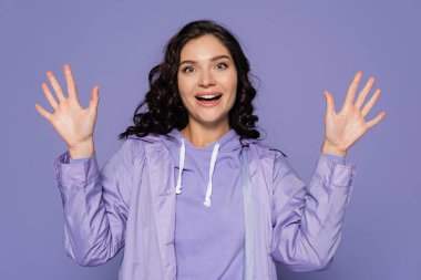 excited young woman in raincoat gesturing isolated on purple clipart