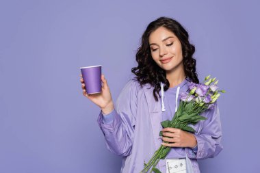 happy young woman in raincoat holding bouquet of flowers and paper cup isolated on purple clipart