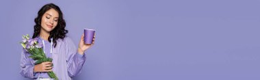 happy young woman in raincoat holding bouquet of flowers and paper cup isolated on purple, banner clipart