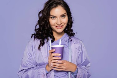 cheerful young woman in raincoat holding paper cup isolated on purple  clipart