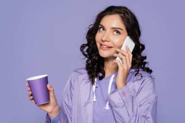 happy young woman in raincoat holding paper cup and talking on smartphone isolated on purple  clipart