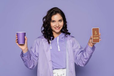 cheerful young woman in raincoat holding paper cup and smartphone with cashback on screen isolated on purple  clipart