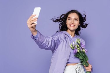 Mor renkte izole edilmiş akıllı telefondan çiçek tutan ve selfie çeken mutlu genç bir kadın.