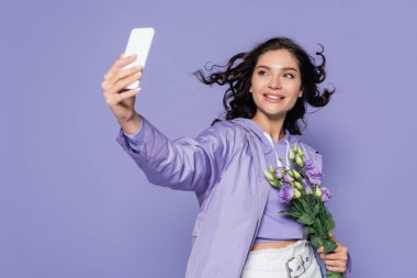 happy young woman holding flowers and taking selfie on smartphone isolated on purple clipart
