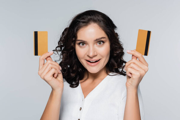 изумленная молодая женщина держит кредитные карты с наличными изолированы на серый