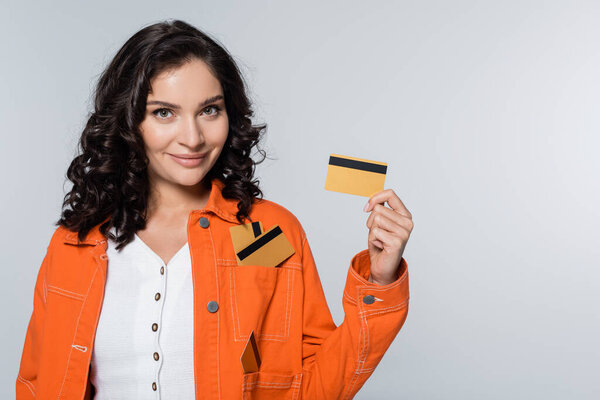 довольная молодая женщина с кредитными картами в кармане оранжевой куртки изолированы на серый