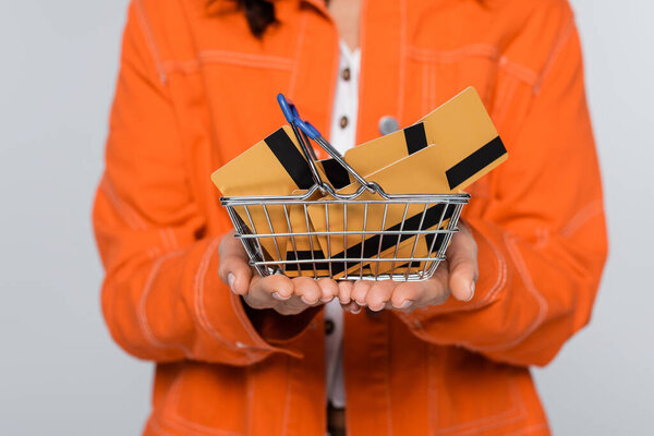 обрезанный вид женщины в оранжевой куртке держит корзину с кредитными картами изолированы на серый